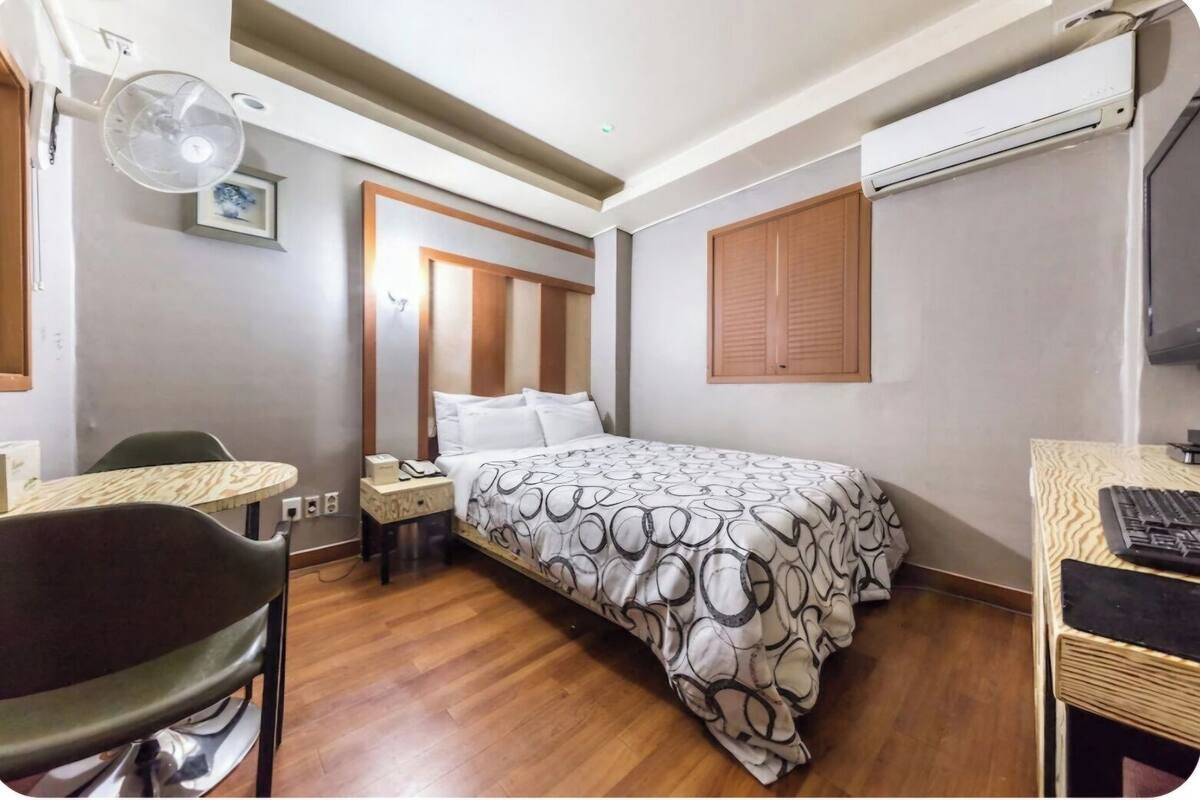 Una habitación de hotel con cama, escritorio con computadora, silla, mesita de noche con lámpara y ventana con persianas.