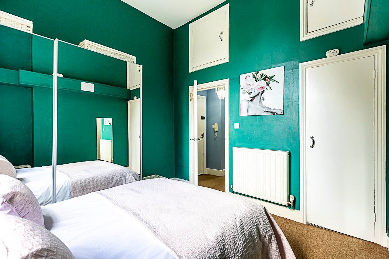 A room with teal walls, two beds with white bedding, a white radiator, and a door with a mirror and a floral painting.