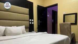 A hotel room with a large bed, a chair, a door leading to another room, and a wall-mounted mirror. - SummerTym Hotel & Suites  (Lekki)
