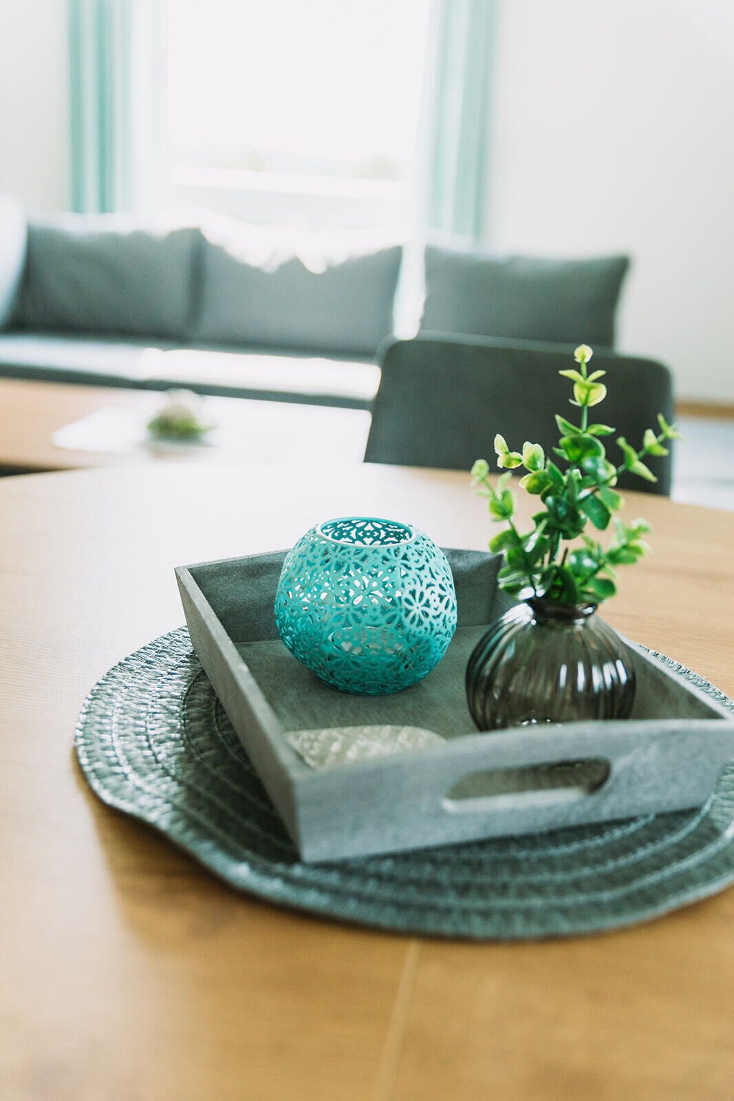 Un plateau orné d’un objet décoratif de couleur turquoise et d’une petite plante, posé sur une table en bois dans une pièce où se trouvent un canapé gris et une fenêtre avec des stores.