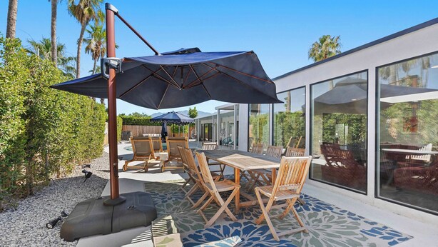 An outdoor patio with a table and chairs, a large umbrella, and a view of the house through large windows. - Aurora Oasis Stylish 4 BDR (Palm Springs)