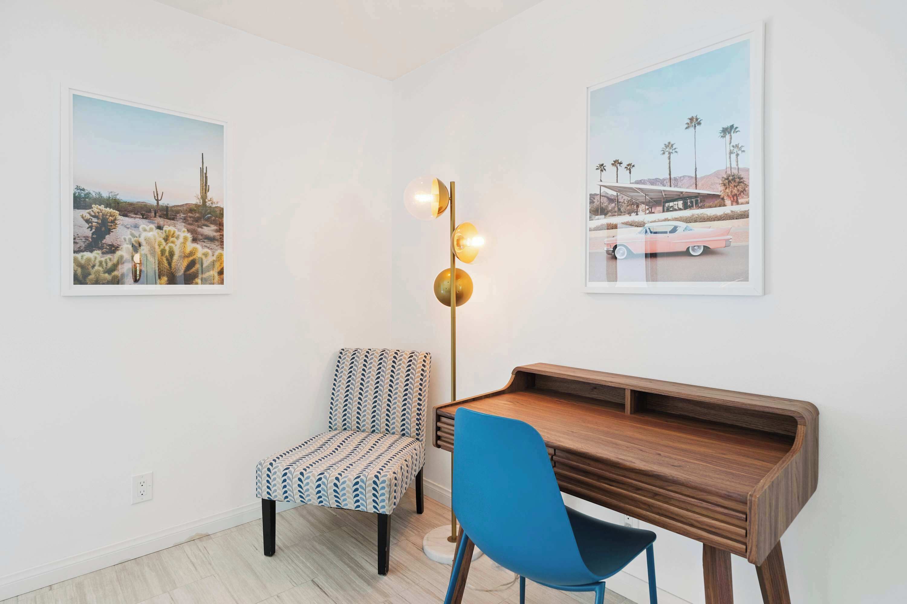 Una habitación con un escritorio de madera, dos sillas, una lámpara de pie y dos cuadros en las paredes.