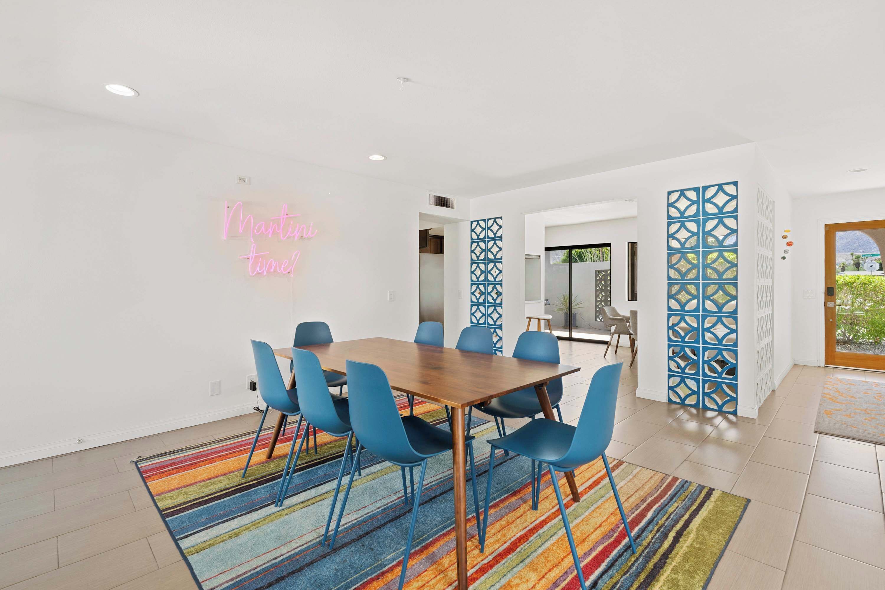 Un comedor moderno con una mesa de madera y sillas azules, un letrero de neón y una alfombra a rayas colorida.