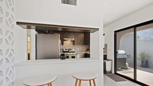 A modern kitchen with a large island, two bar stools, and a sliding glass door leading to an outdoor patio. - Aurora Oasis Stylish 4 BDR (Palm Springs)
