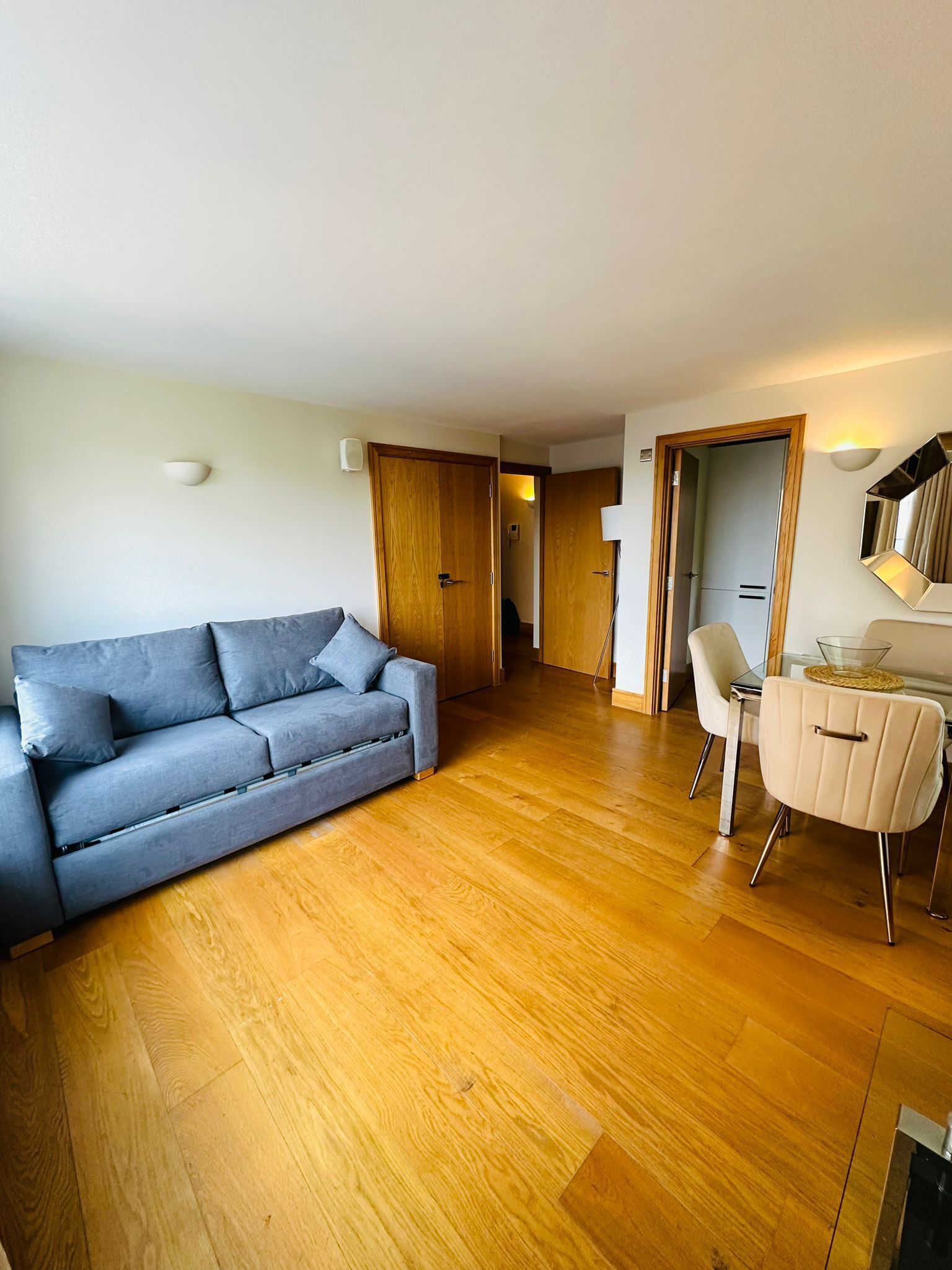A modern living room with a grey sofa, a dining table with chairs, wooden flooring, and a wooden door.