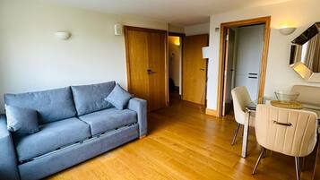 A modern living room with a grey sofa, a dining table with chairs, wooden flooring, and a wooden door.