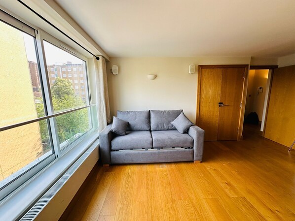 Una habitación con un sofá gris, suelo de madera, una ventana con persianas y una puerta.