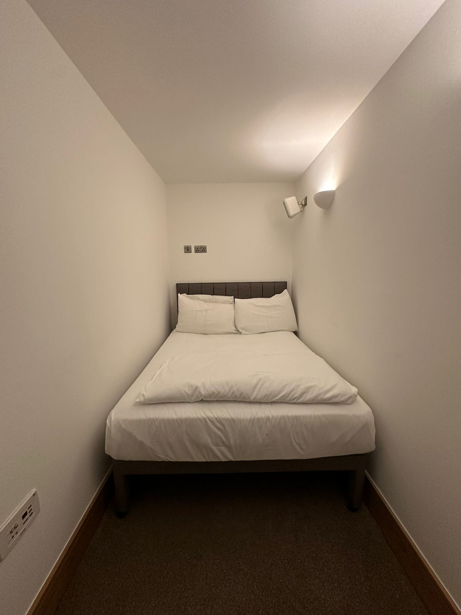 A small, narrow hotel room with a single bed, a nightstand, and a wall-mounted light fixture.