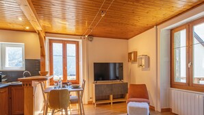 A cozy living room with a wooden ceiling, a TV, a dining area with chairs, and a kitchen visible through an open door. - Rimaye Blanche - A bright and cozy 1 bed apartment. (Chamonix-Mont-Blanc)