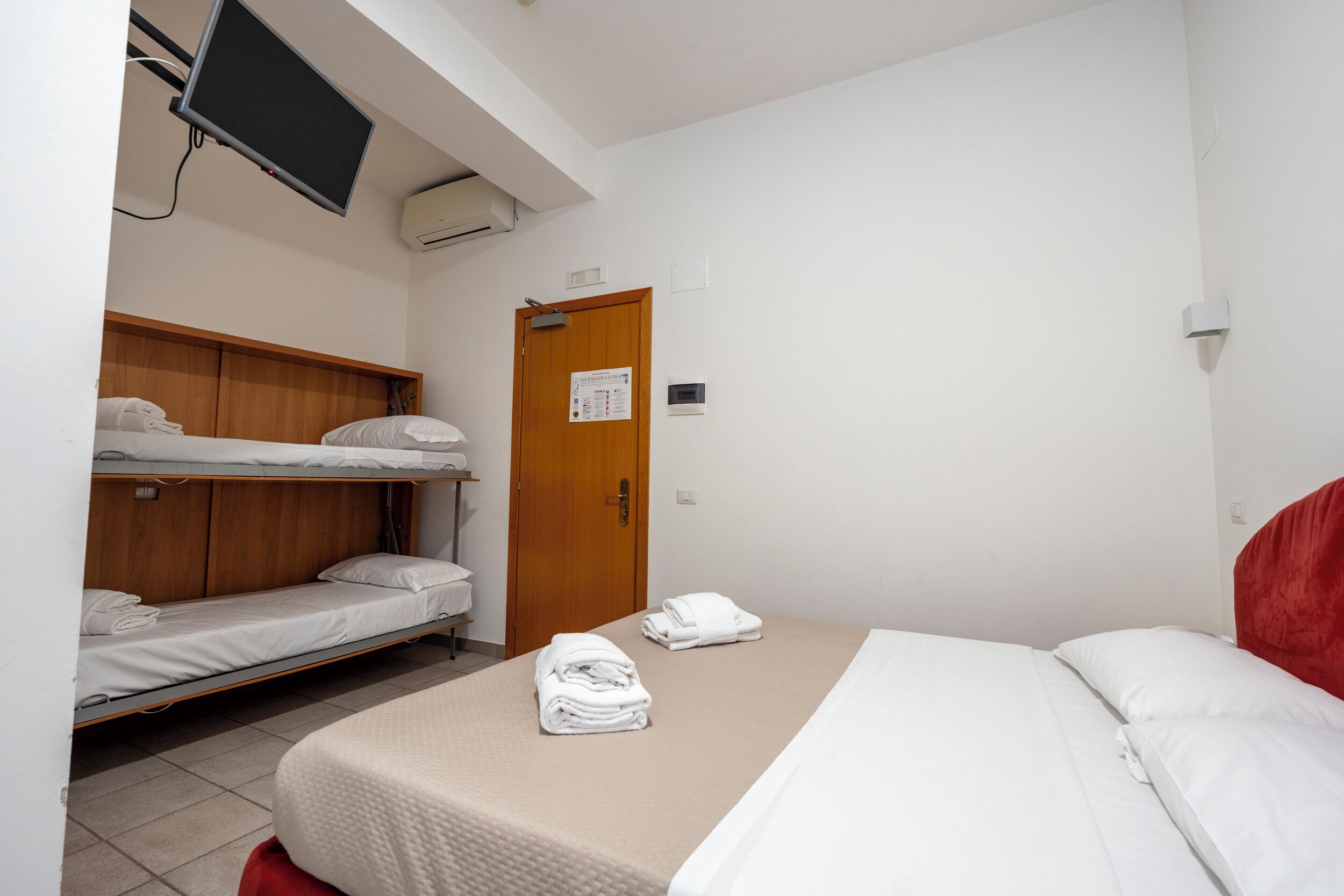 Quarto de hotel com uma cama de solteiro, uma cama beliche, uma televisão e uma porta com um aviso.