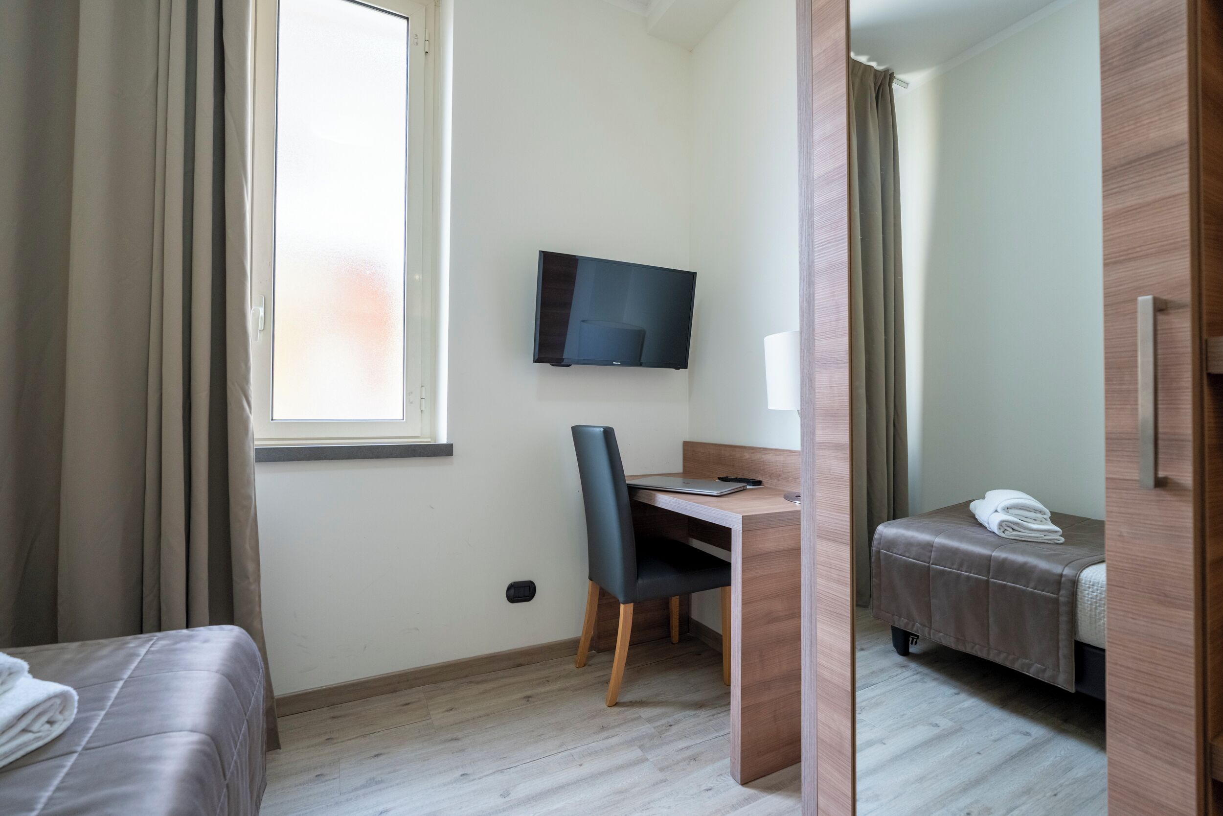 A hotel room with a bed, a desk with a chair, a television mounted on the wall, and a window with curtains.