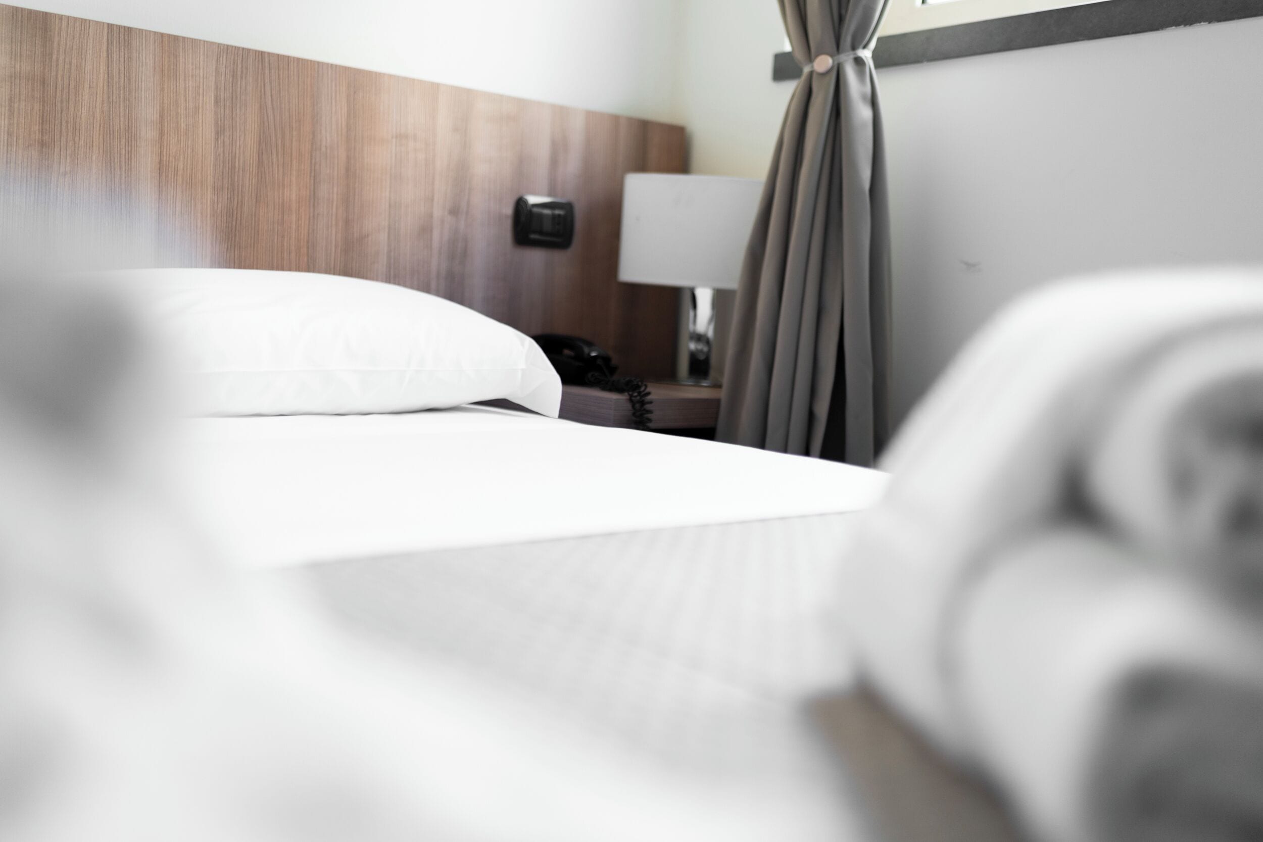 Quarto de hotel com cabeceira de madeira, cama com roupa de cama branca, abajur e telefone.