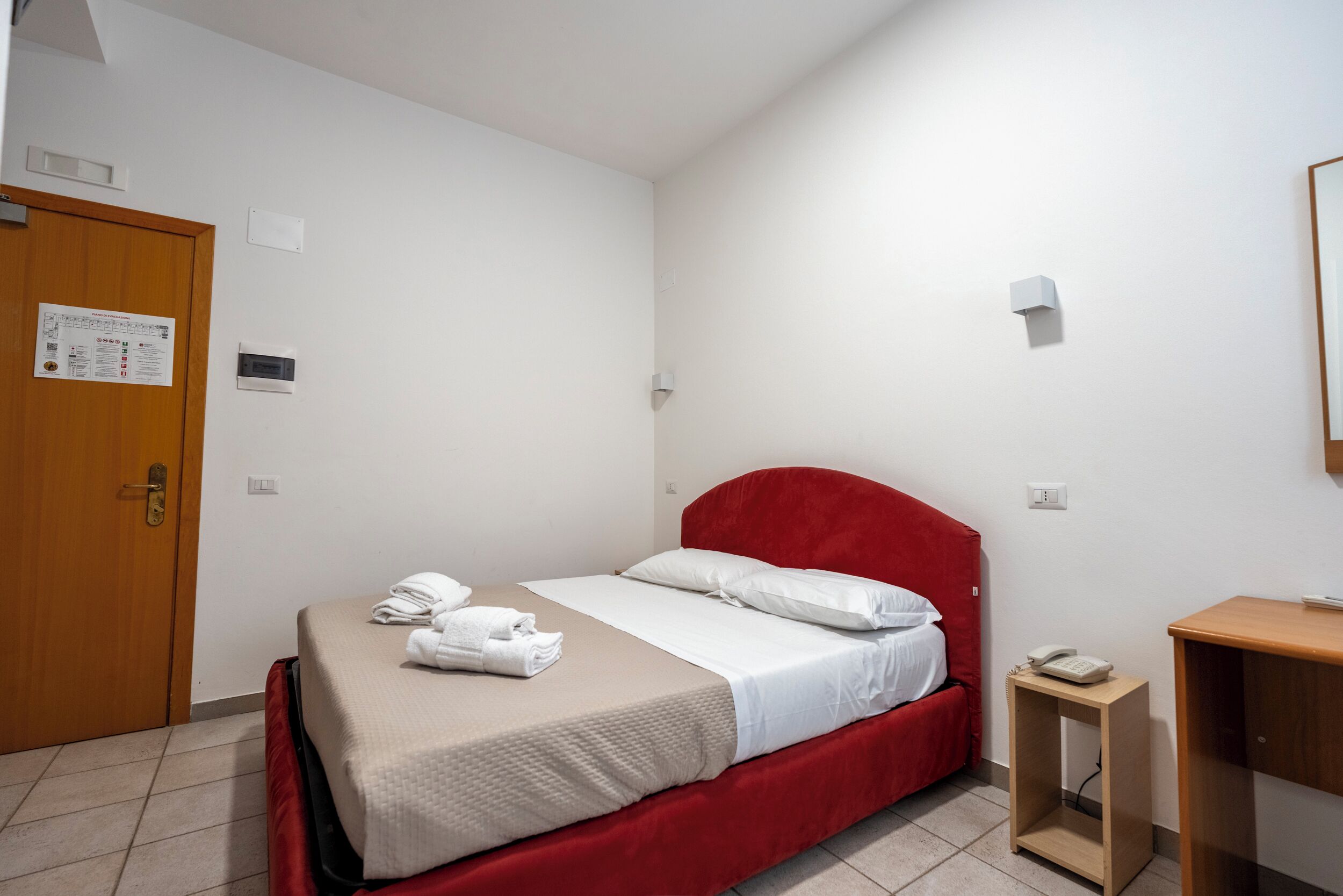Una habitación de hotel con cama, una puerta de madera, una mesita de noche pequeña con un teléfono y una lámpara fijada a la pared.