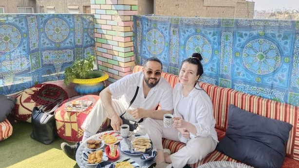 Una pareja sentada en un sofá a rayas, con un muro estampado de fondo, una mesa con comida y bebidas, y un pilar de ladrillo.