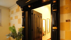 Un vano con marco de madera oscura y detalles dorados, que da acceso a una habitación con suelo a cuadros y un espejo en la pared.