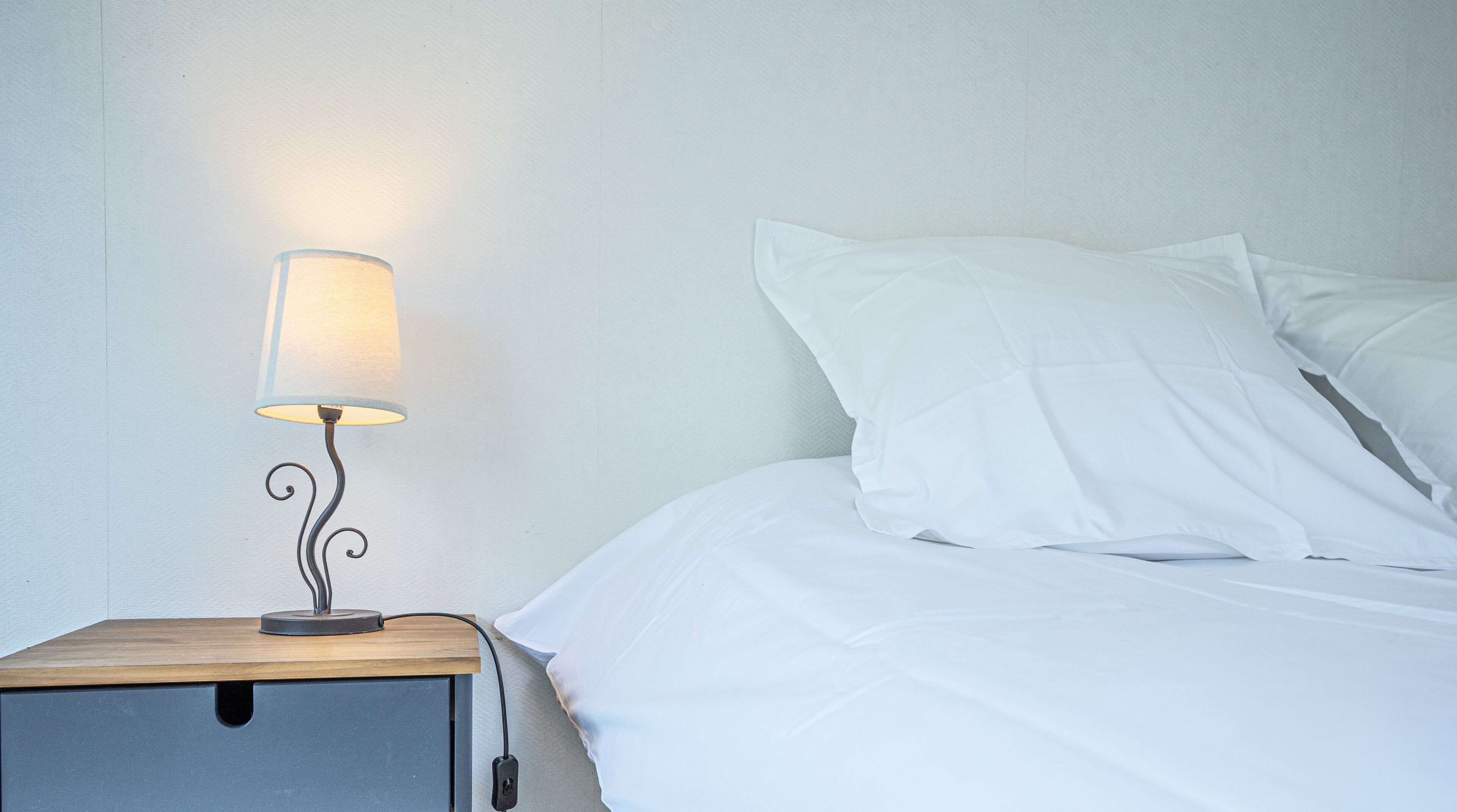 Una cama bien hecha con sábanas blancas, una mesita de noche con una lámpara y una pared con textura.