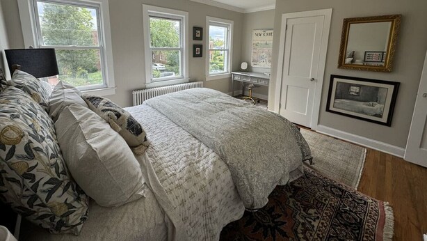 A bedroom with a large bed, a desk, and a chair, a radiator, and a window with a view of greenery. - Old Town Gem – Steps to Cafés, Shops & Metro! (Alexandria)