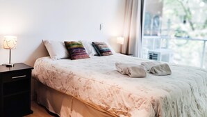 Een net opgemaakt bed met bloemenbeddengoed en meerdere kussens, een nachtkastje met een lamp, en uitzicht op groen door een raam.
