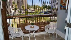 A balcony with two white chairs and a small round table, overlooking a street with palm trees and parked cars. - Relaxing Studio Near El Yunque With Pool View (Luquillo)