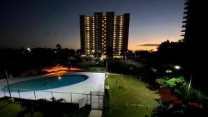 A high-rise building with illuminated windows at dusk, a swimming pool with a fenced area, and a grassy lawn with a small structure. - Relaxing Studio Near El Yunque With Pool View (Luquillo)