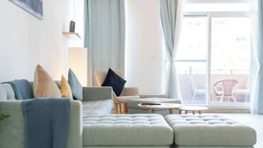 Una sala moderna con un sofá modular, una mesita redonda y una mesita auxiliar pequeña. Se ve un balcón con mobiliario exterior a través de la ventana.