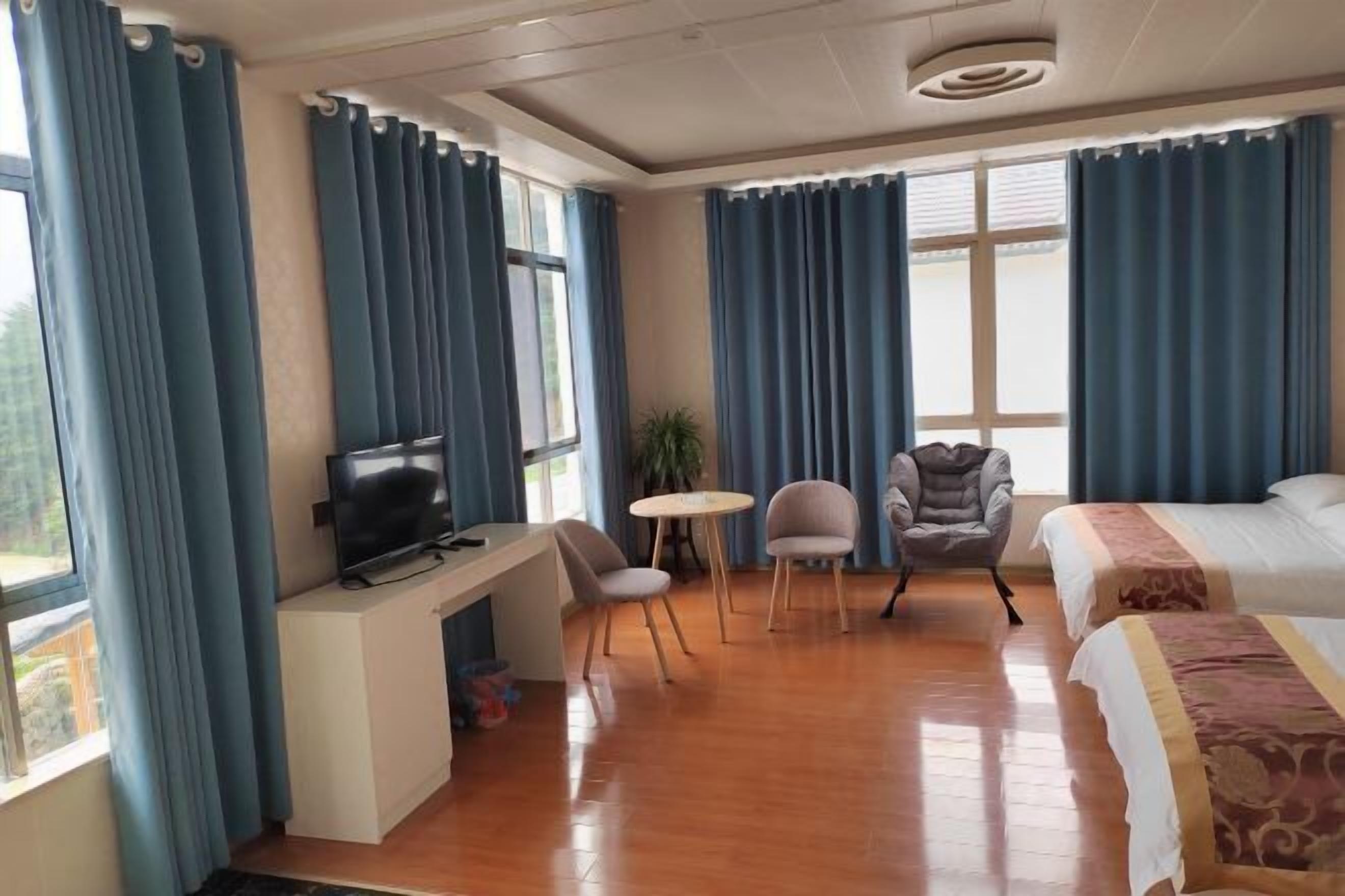 Habitación de hotel con televisión, dos sillas, una mesita, cama y ventanas amplias con cortinas.