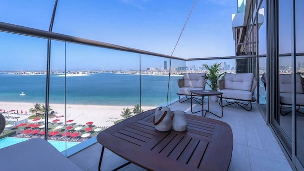 A balcony with a view of the beach and city skyline, furnished with a table, chairs, and a small plant. - StayShort - Cozy 2BR in Palm Jumeirah with Beach Views! (Palm Jumeirah)