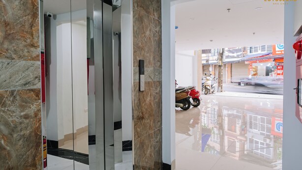 A modern hotel lobby with a marble floor, a glass elevator, and a view of the street outside. - Lotus Land Apartment (Hanoi)