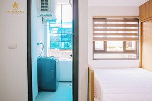 Een hotelkamer met een bed, een raam met zonwering, een wasmachine en uitzicht op een blauw gebouw.