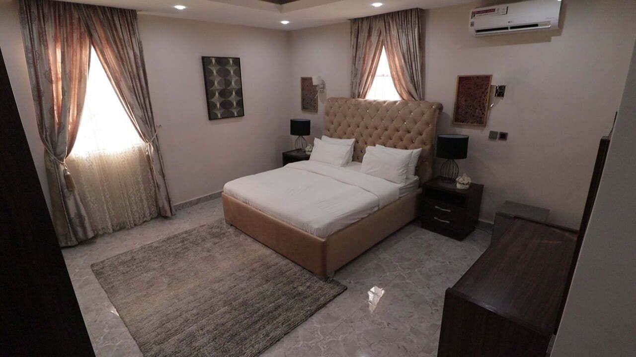 A bedroom with a large bed, two bedside tables, a nightstand, a rug, and an air conditioning unit on the wall.