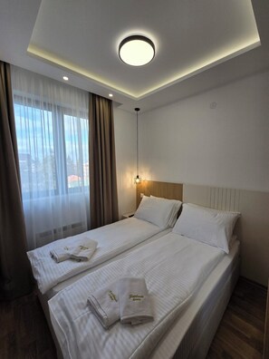 Ein Hotelzimmer mit zwei Betten, einem großen Fenster mit durchsichtigen Vorhängen, einer Deckenleuchte und einem Nachttisch mit einer Lampe.