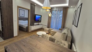 Un salon moderne avec une table à manger en bois, un ensemble de canapés, un téléviseur fixé sur un panneau en bois et une grande fenêtre avec des rideaux.