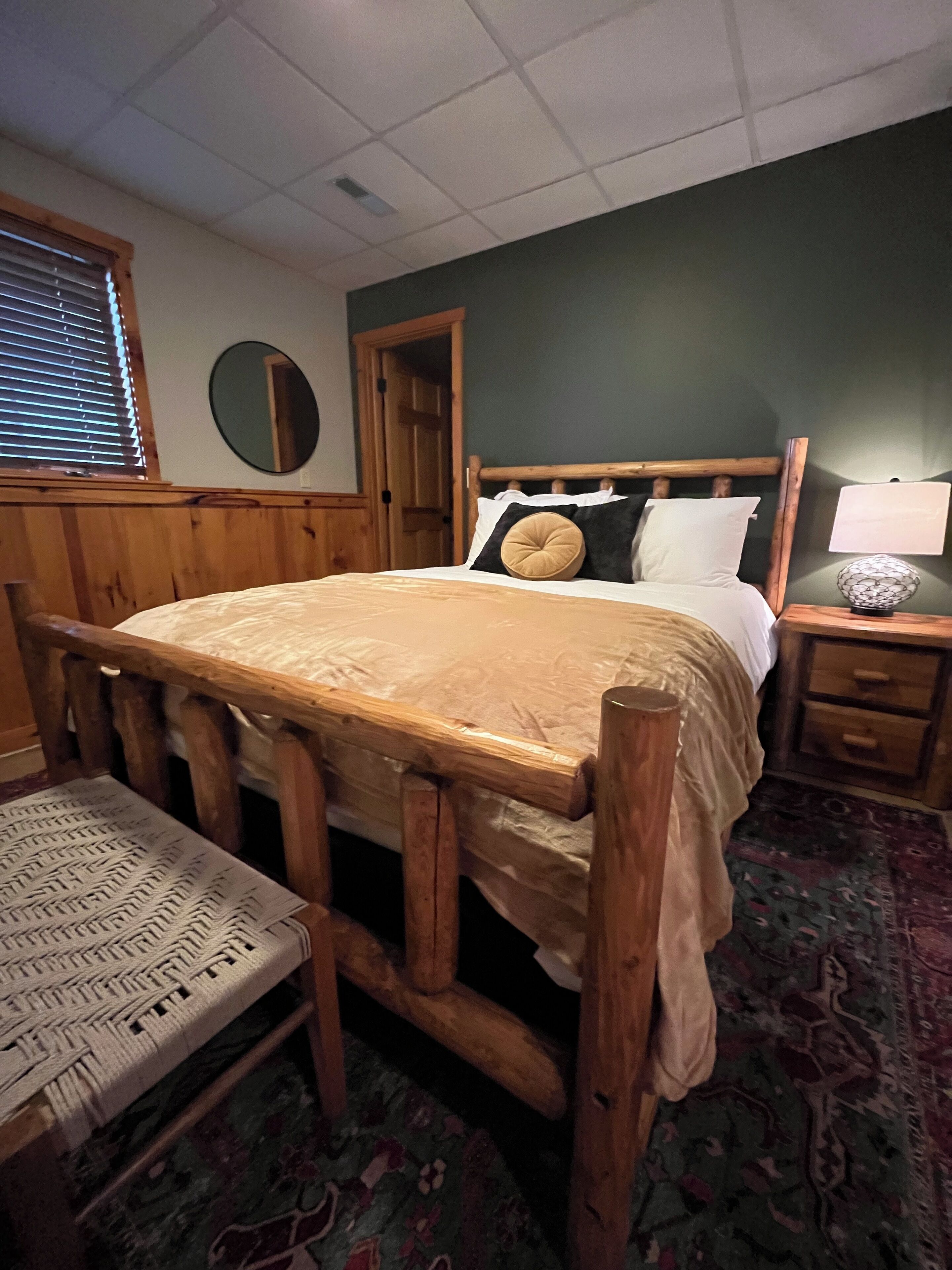 Una cama con estructura de madera, cubierta con un edredón blanco y una almohada marrón. A la derecha, una mesita de noche de madera con una lámpara y un cajón pequeño. Un banco tejido está frente a la cama.