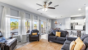 A spacious living room with a large window, a ceiling fan, and a kitchen area visible through an open door. - Romar Lakes 103B w Pool (Orange Beach)