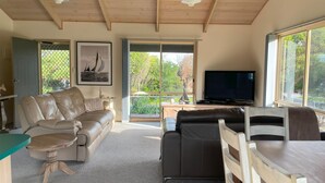 Amplia sala de estar con un sofá de cuero color beige, un sofá de cuero negro y una mesa de comedor de madera. Hay un ventanal con vistas a árboles y un velero. Un televisor está montado en la pared.
