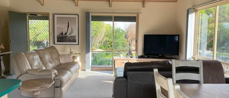 Amplia sala de estar con un sofá de cuero color beige, un sofá de cuero negro y una mesa de comedor de madera. Hay un ventanal con vistas a árboles y un velero. Un televisor está montado en la pared.