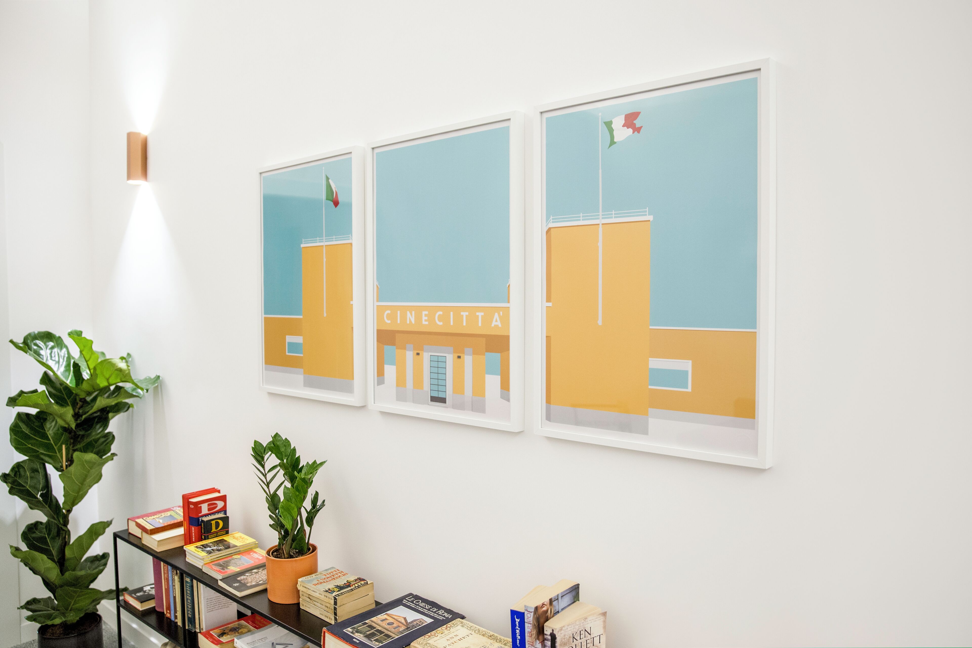 Una pared con tres cuadros enmarcados de un edificio que muestra el letrero 'CINECITTA'. Hay libros en una estantería y dos plantas en macetas.