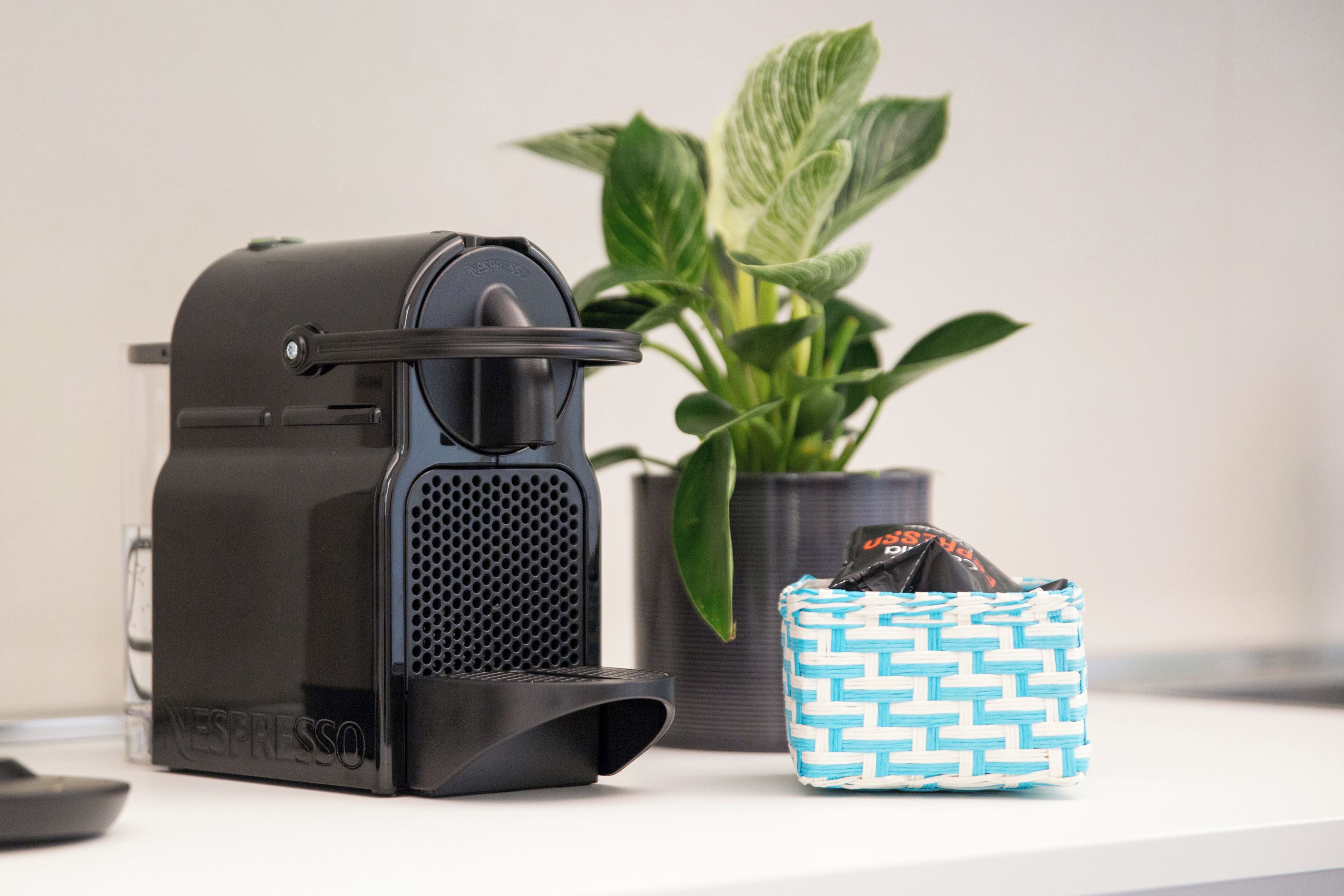 Une machine à café Nespresso, un sac en tissu tressé et une plante en pot, le tout posé sur une surface blanche.