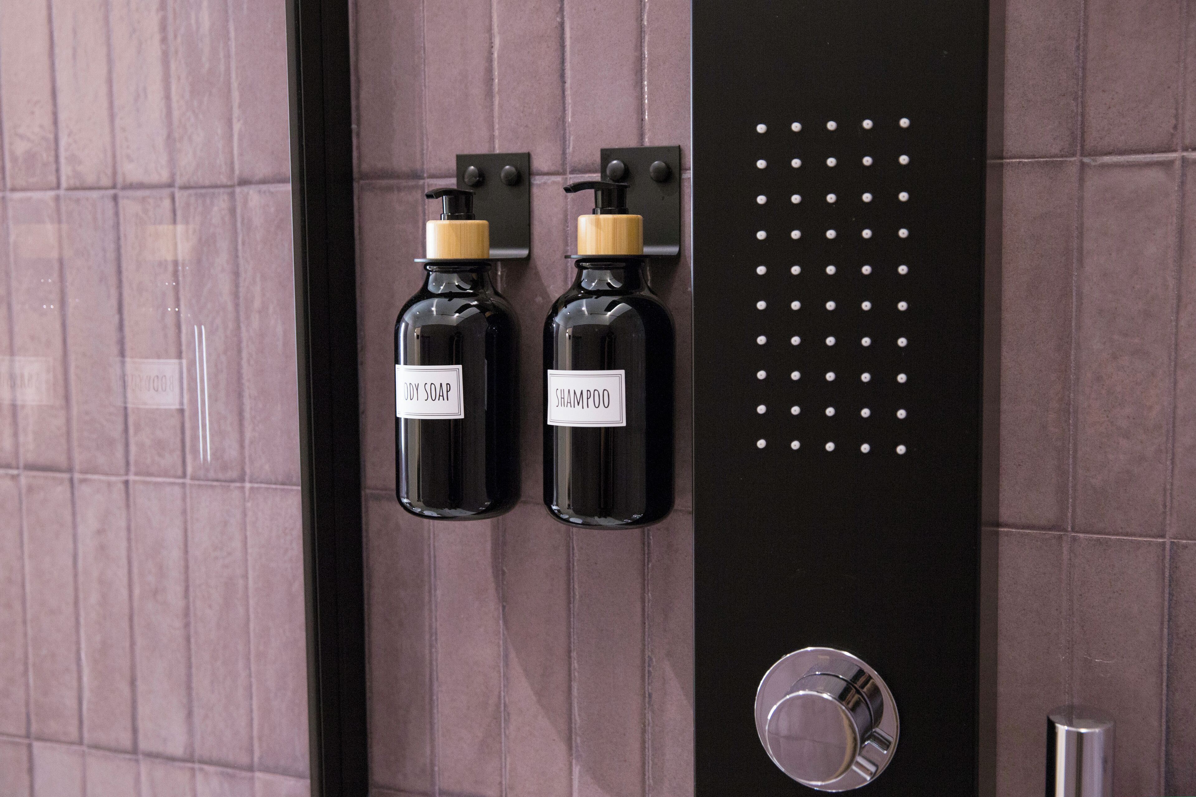 A bathroom with two black bottles labeled "Dry Soap" and "Shampoo" mounted on a wall with a metallic panel and a round faucet.