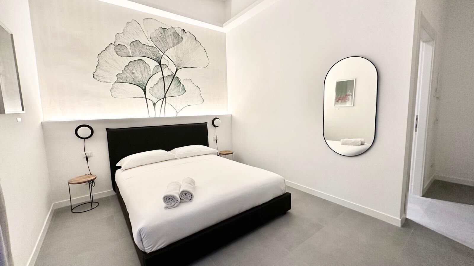 A modern hotel room with a large bed, a round mirror, and a wall art piece.