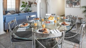 Een eetruimte met een glazen tafel, omringd door metalen stoelen, met ontbijtproducten zoals croissants, fruit en sap.