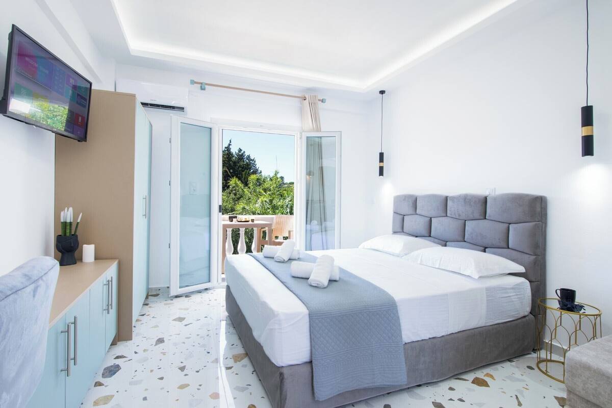 A modern bedroom with a large bed, a TV mounted on the wall, a balcony with a view of greenery, and a small table with a cup.