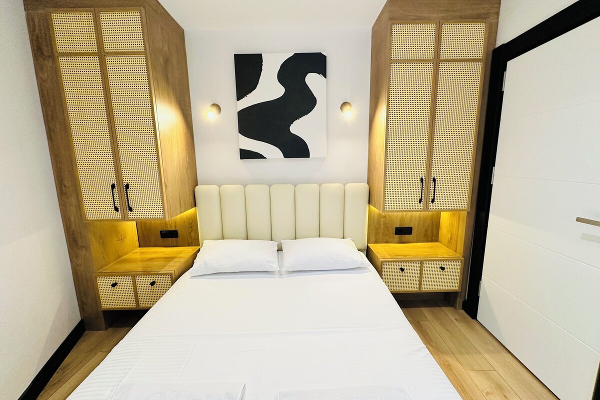 A modern hotel room with a large bed, wooden headboard, and a wall-mounted abstract art piece.