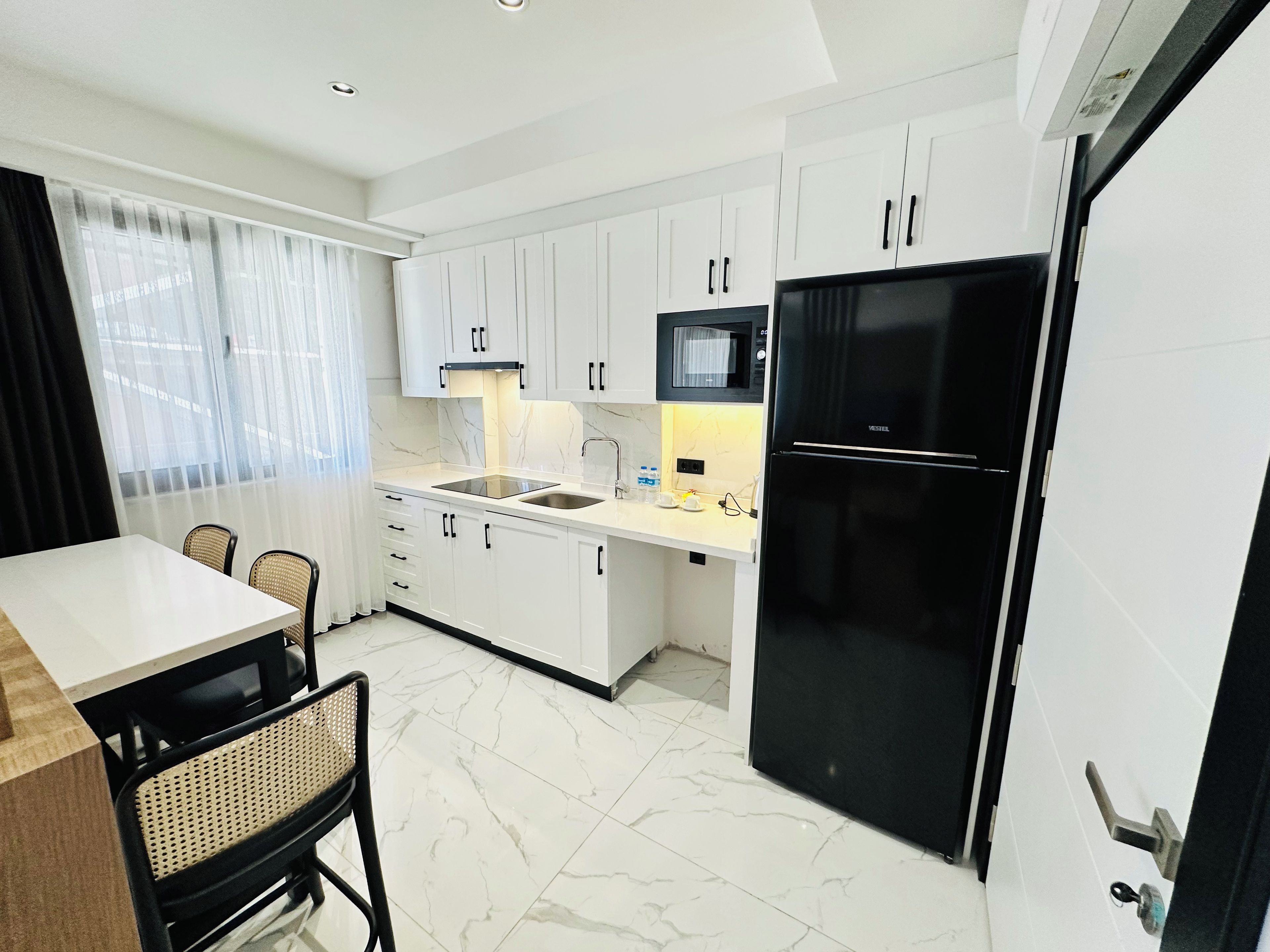 Una cocina moderna con armarios blancos, un refrigerador negro y una zona de comedor con mesa y sillas.