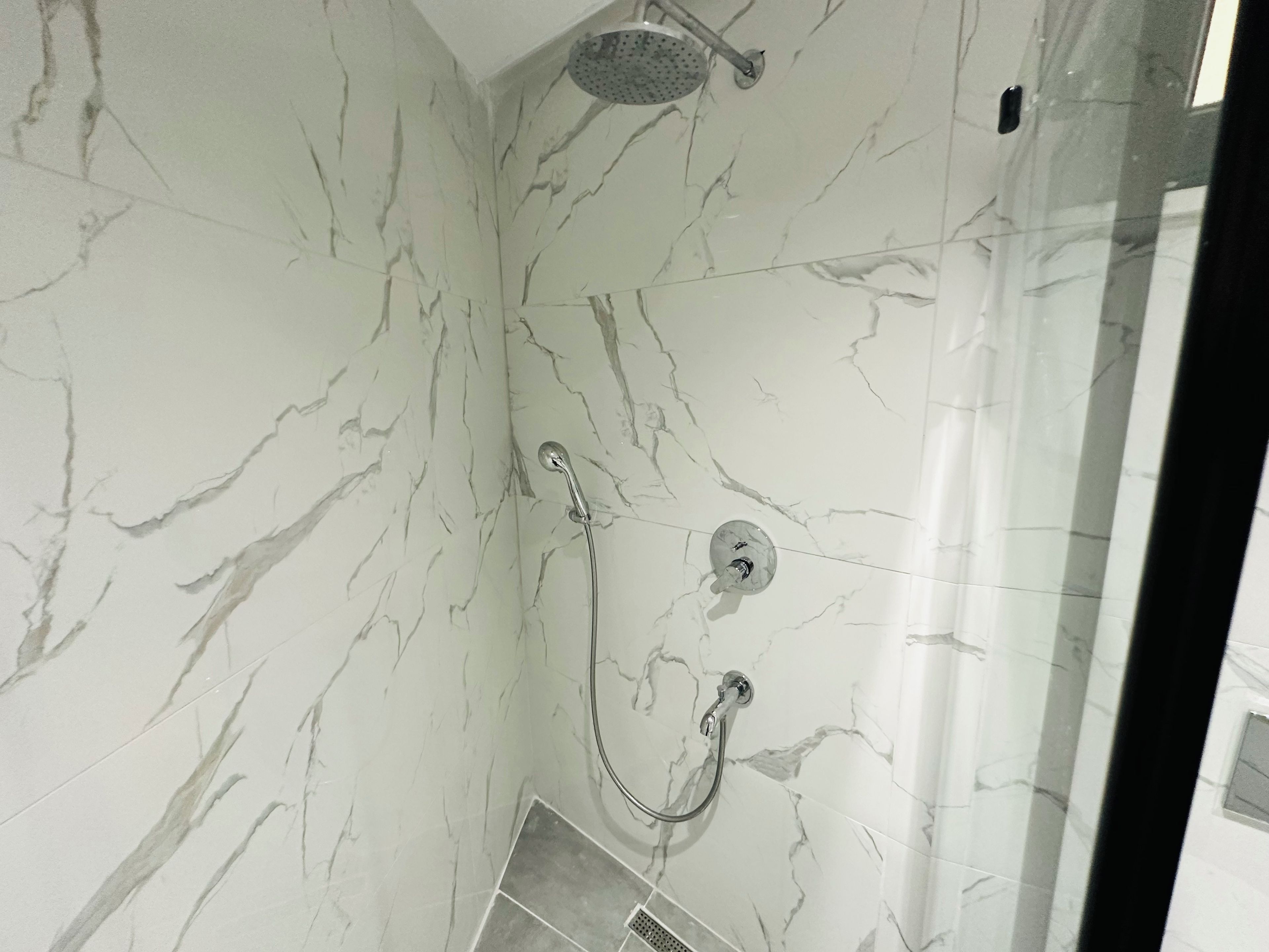 Un espacio de ducha con mampara de vidrio, paredes de mármol y un cabezal de ducha manual.