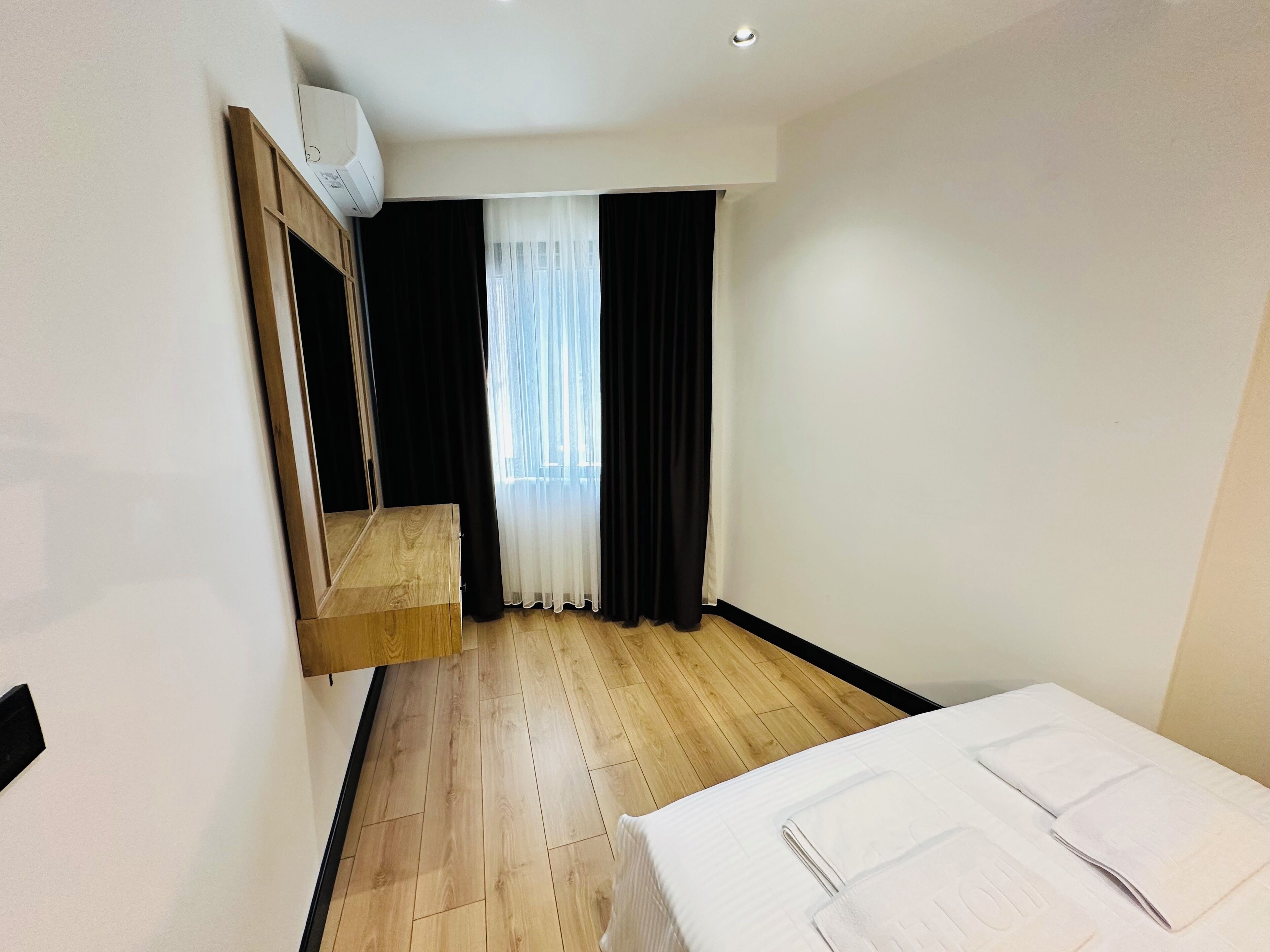 A hotel room with a wooden floor, a bed with white linens, a built-in wardrobe, a window with sheer curtains, and a ceiling-mounted air conditioning unit.