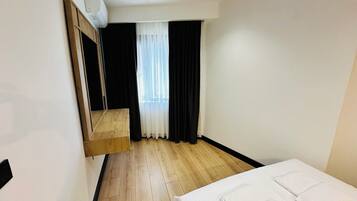 A hotel room with a wooden floor, a bed with white linens, a built-in wardrobe, a window with sheer curtains, and a ceiling-mounted air conditioning unit.