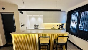 A modern kitchen with a central island, two bar stools, a coffee machine, and a window with blinds. - Rose Point Residence (Istanbul)