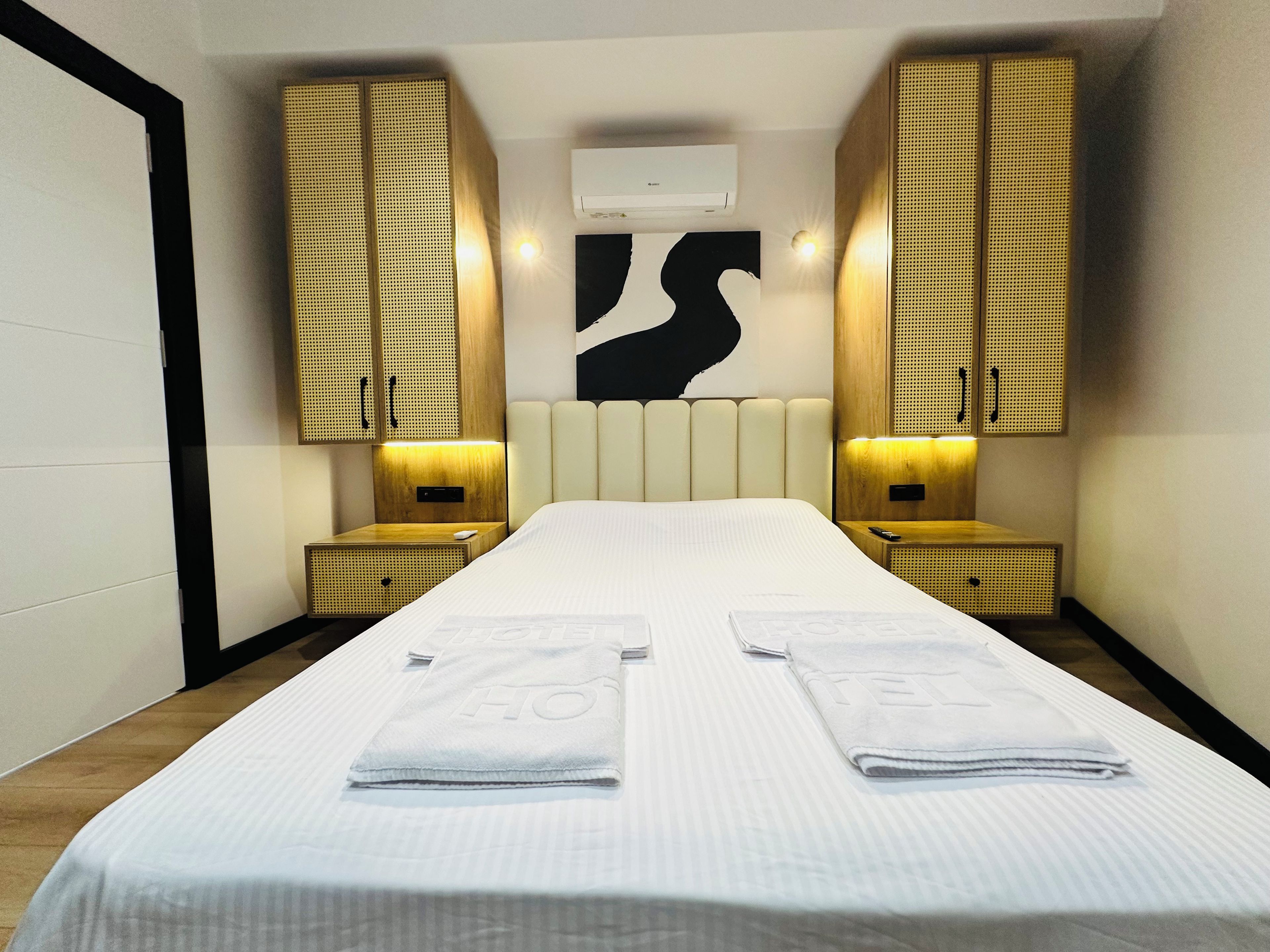 Une chambre d’hôtel dotée d’un grand lit, de deux tables de chevet, d’un climatiseur mural et d’une œuvre d’art murale originale.