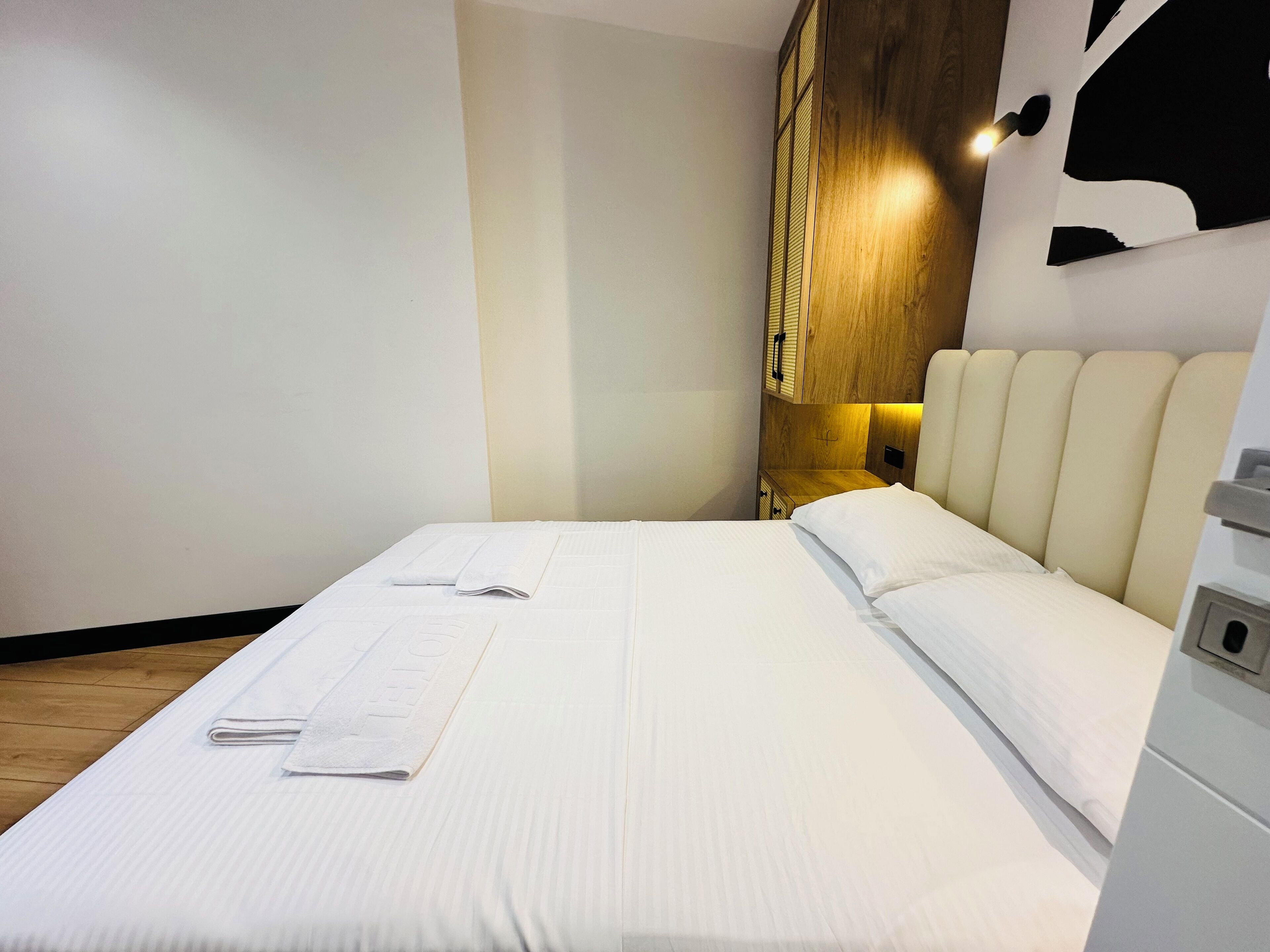 A neatly made bed with white linens and two folded towels, a wooden headboard with a light fixture, and a small wooden nightstand with drawers.
